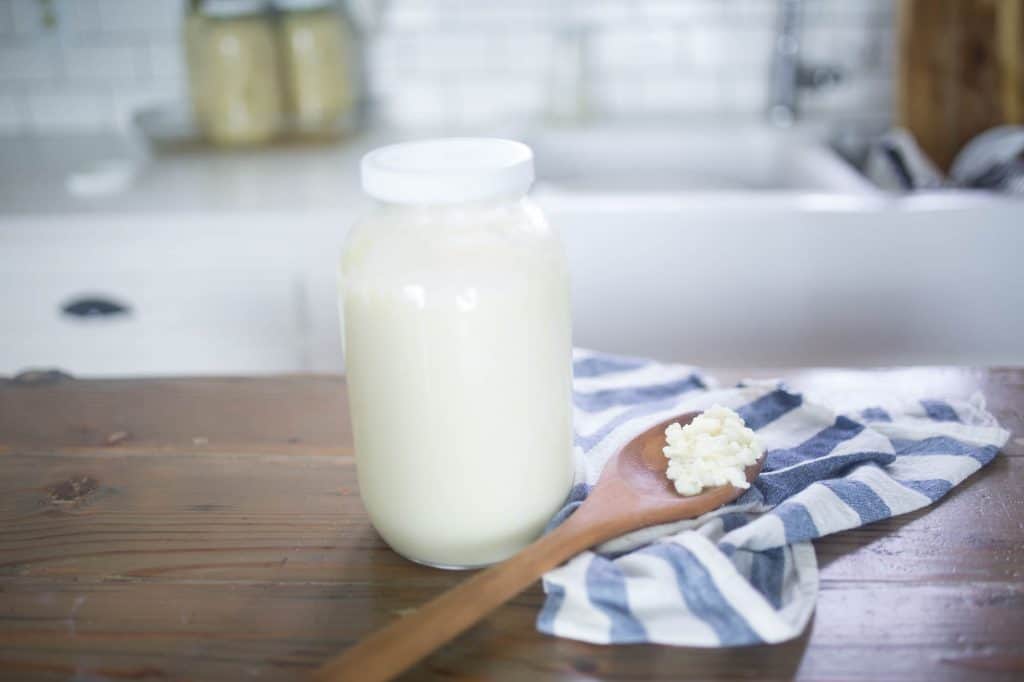 homemade milk kefit in a half gallon mason jar with a white lid on a wood contertop. A white and blue stripped towel with a wood spoon full of kefir grains sits on top of the towel.