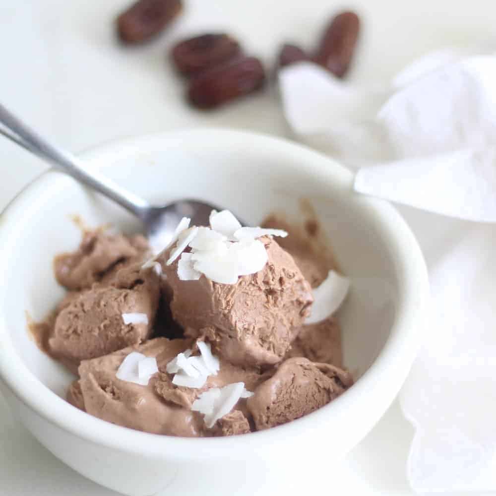 homemade chocolate ice cream topped with coconut chips in a white bowl with a spoon in the bowl. A white napkin is to the right with dates behind