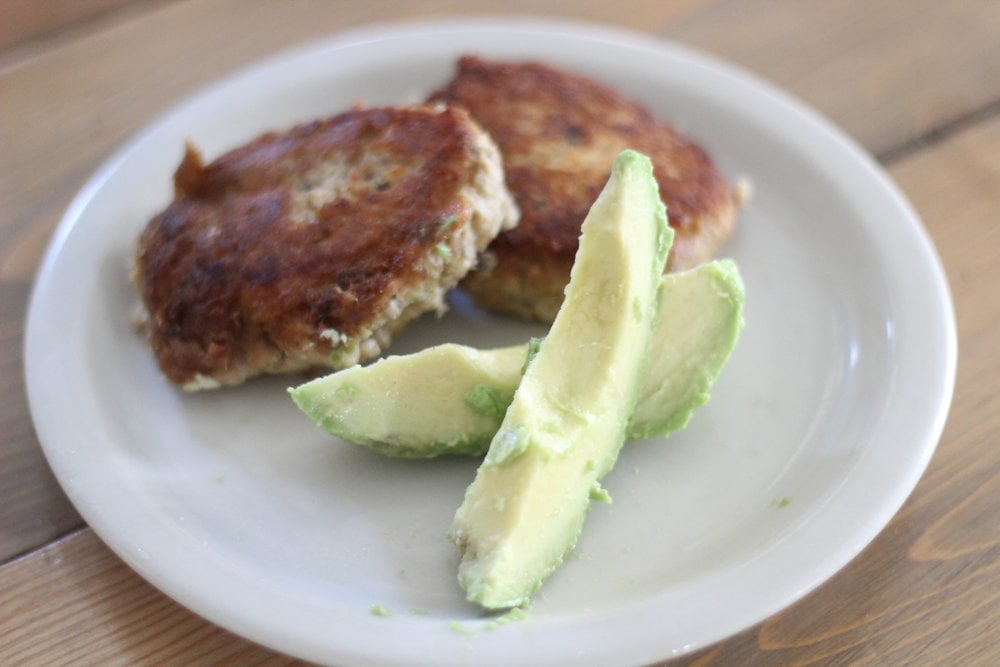 two crispy salmon burgers on a white plate with sliced avocado