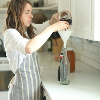 woman adding grape juice into a flip top bottle with a funnel in a white kitchen