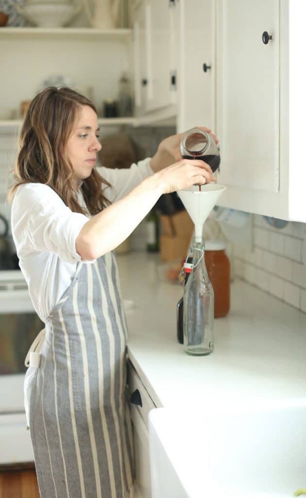 woman adding grape juice into a flip top bottle with a funnel in a white kitchen