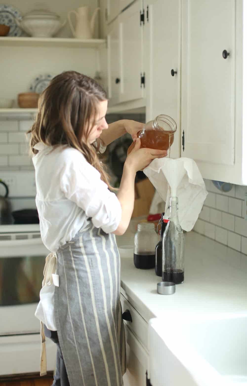 women wearing a white shirt and a stripped apron is pouring a mason jar full of water kefir into a bottle