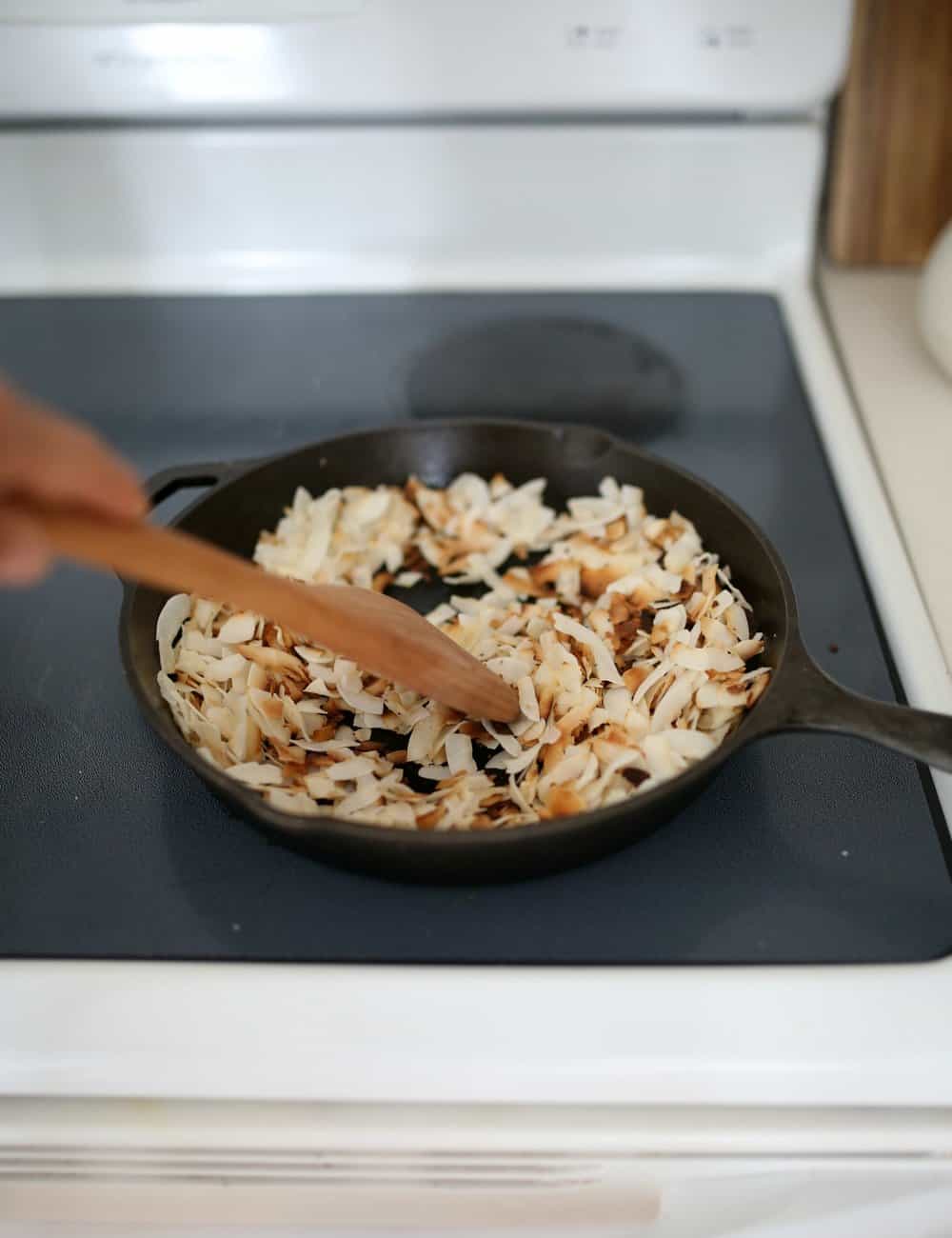 coconut flakes being toasted o a cast iron skillet with a wooden spoon.