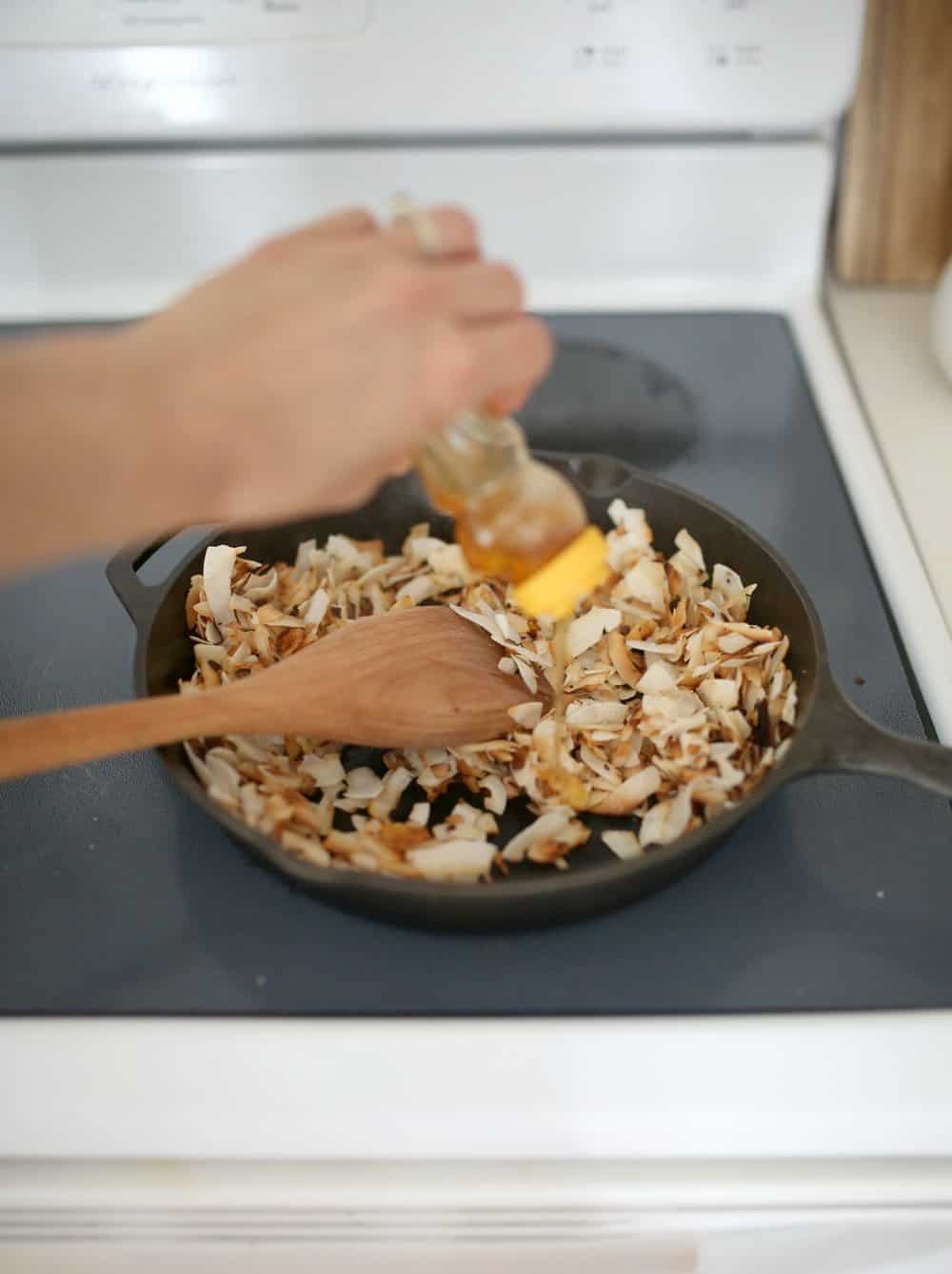 adding honey to toasted coconut chips in a cast iron skillet