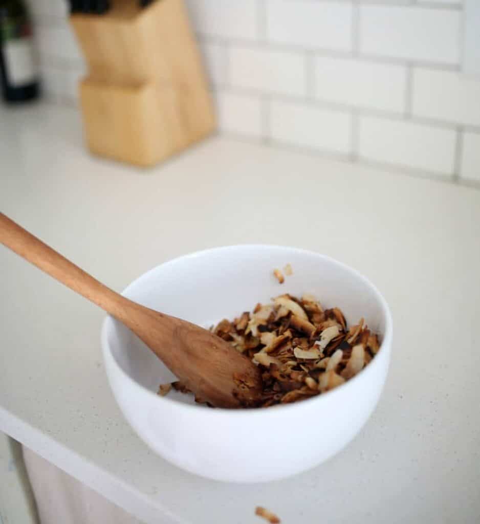 Toasted coconut chips in a white bowl with a wooden spoon on a white quartz countertop