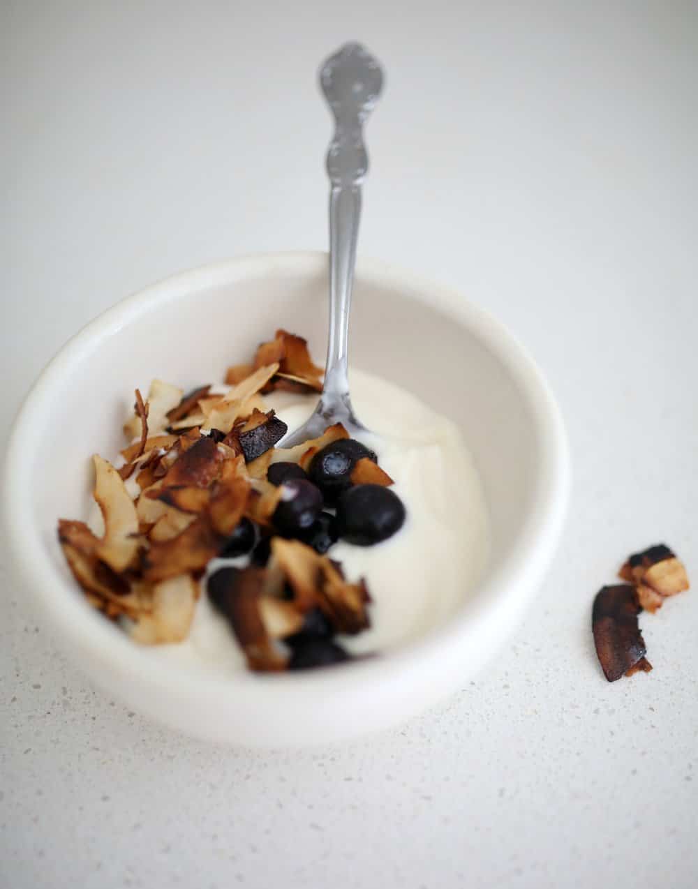 yogurt topped with toasted coconut chips and blueberries in a white bowl with a metal spoon