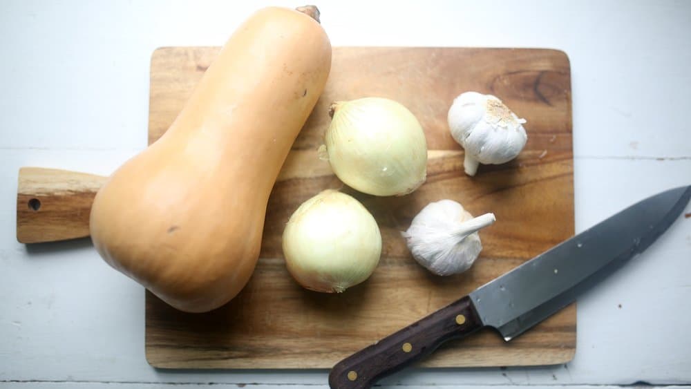 butternut squash, two onions, and two head of garlic on a cutting board with a knife - roasted butternut squash soup