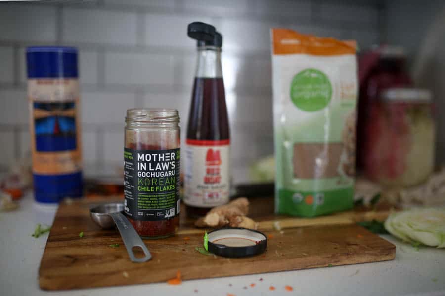 homemade kimchi ingredients including: Korean chili flakes, fish sauce, brown sugar, fresh ginger, on a wooden cutting board in a white kitchen.