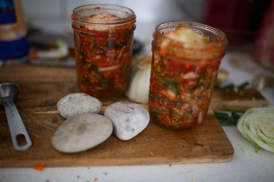 two pint mason jars of homemade kimchi on a wood cutting board next to some clean rocks to use as weights