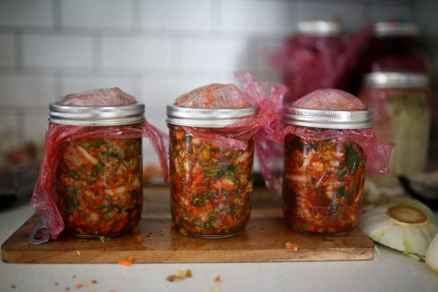 three jars of kimchi topped with plastic wrap and mason jar rinks on a wood cutting board.
