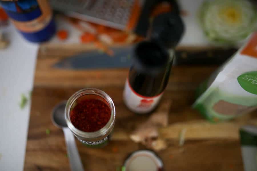 overhead photo of Korean chili flakes in a glass dish on a wooden cutting board. A container of fish sauce, some fresh ginger, and a package of brown sugar also sit next to the chili flakes on a wood cutting board.