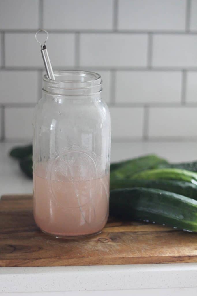 large mason jar filled with a salt water brine on a cutting board next to cucumbers