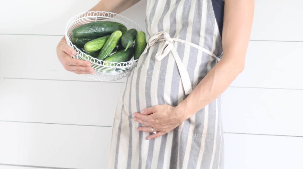 women in a stripped apron holding a basket of cucumbers with one hand and holding her pregnant belly with another hand.
