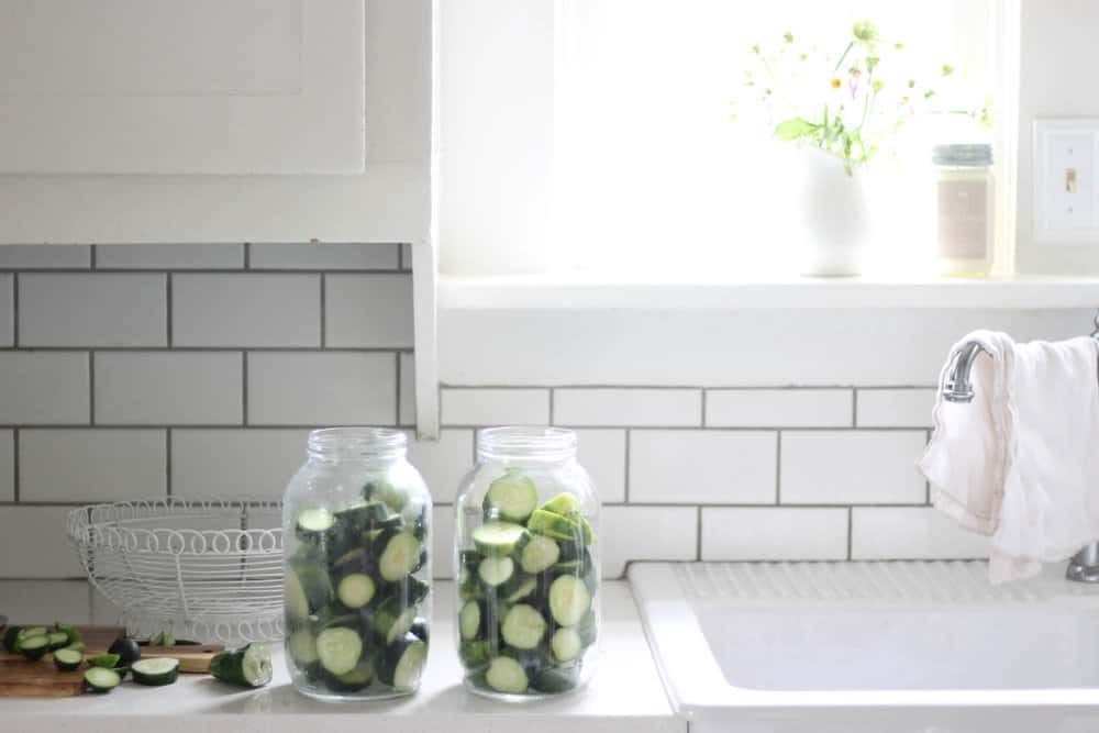 sliced cucumbers in half gallon mason jars on a white kitchen countertop next to a farmhouse sink