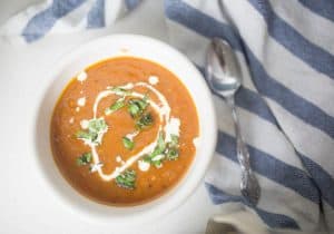 Recipe for roasted red pepper and tomato soup in a white bowl on a blue and white stripped towel with a spoon to the right