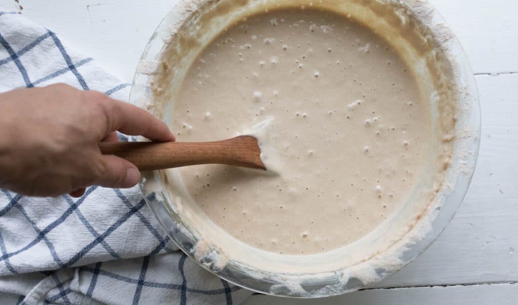 hand holding a wooden spoon in a bowl of sourdough starter with a blue and white towel to the left