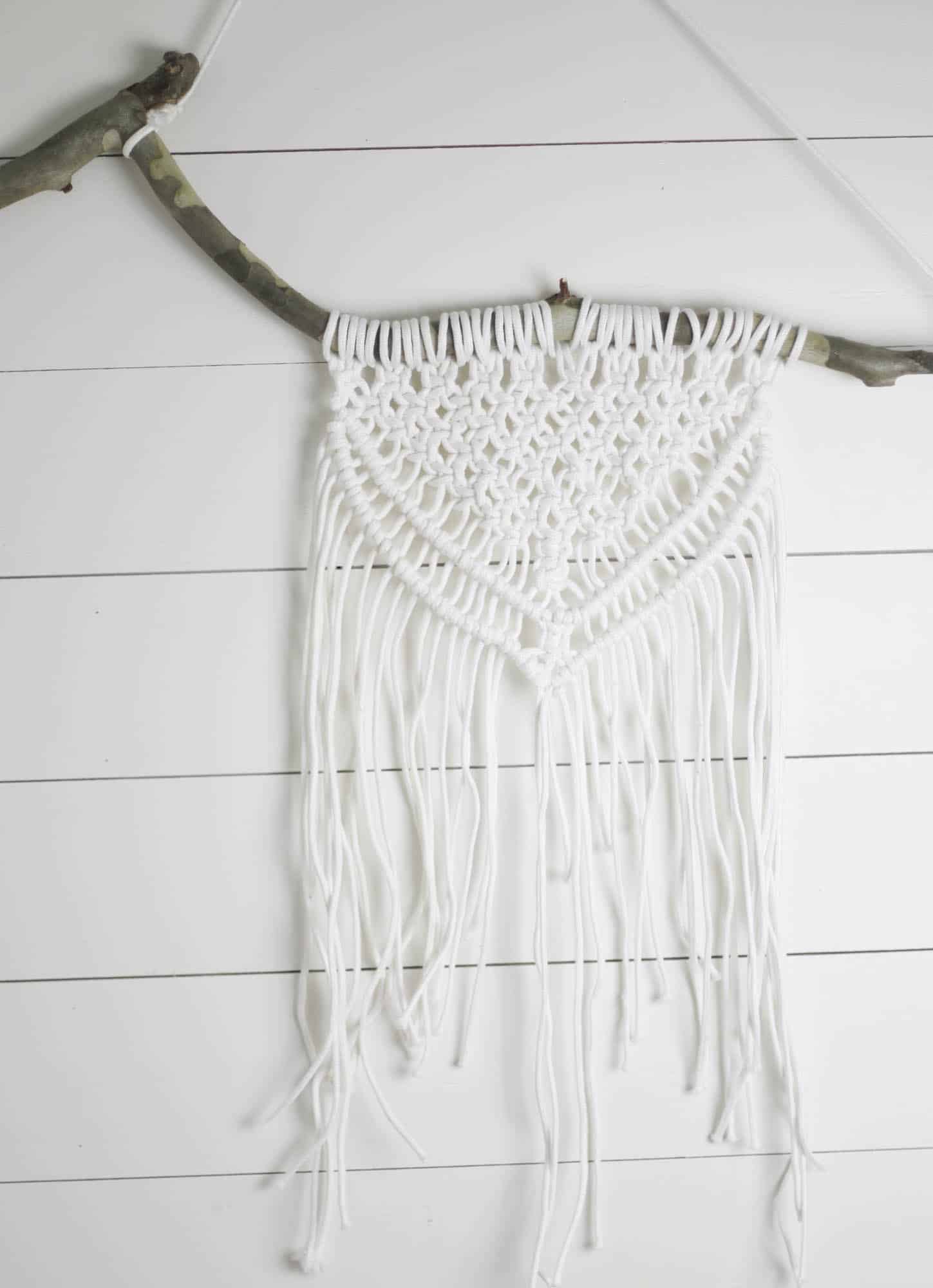 a piece of macrame wall decor on that hangs on a branch against a white shiplap all