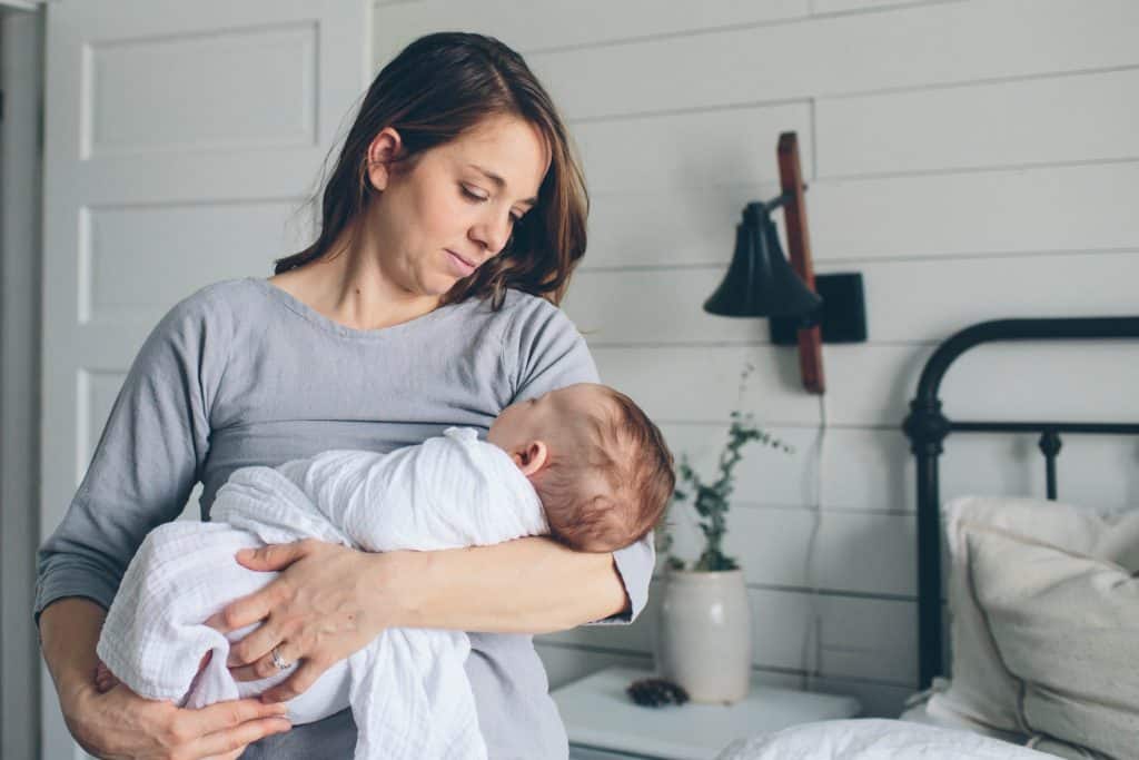 mom wearing a gray dress looking at her baby in her arms with a muslin blanket wrapped around him