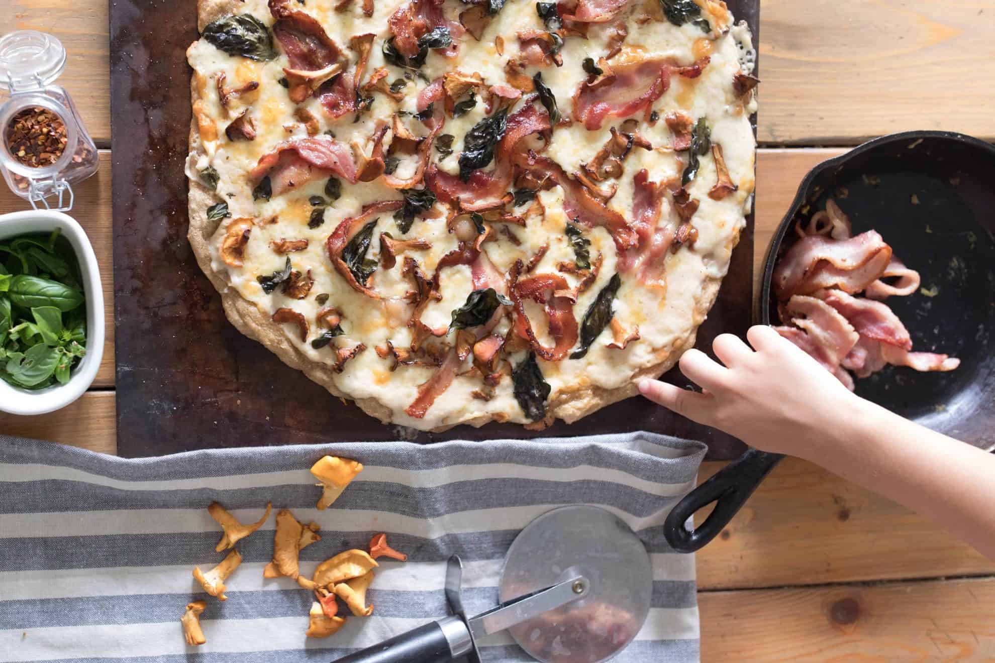 overhead shot of bacon chanterelle mushroom pizza on a pizza stone with ingredients surrounding itforaging chantrelle mushrooms wild edibles