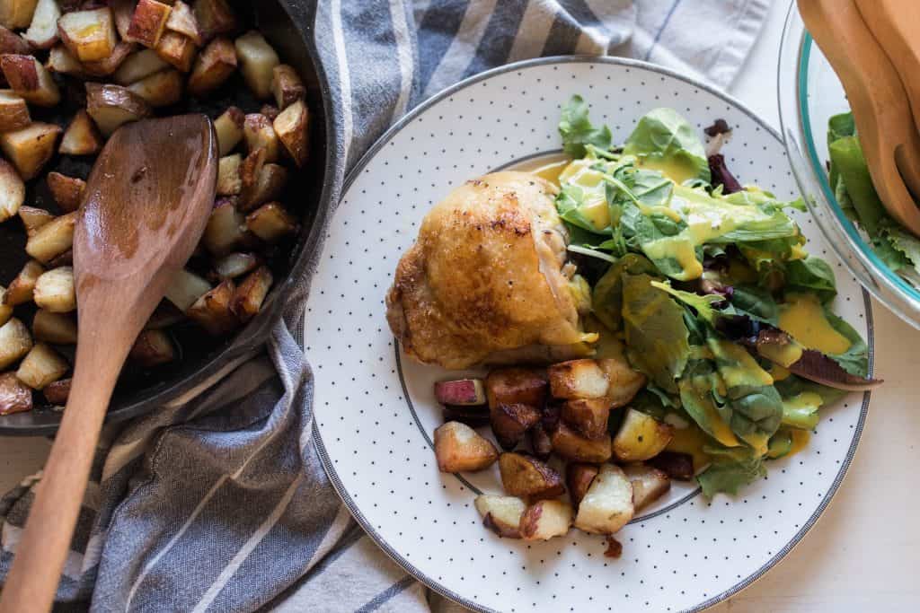 Lemon Rosemary Chicken with potatoes and salad on a white and gray plate. A skillet of potatoes to the left.