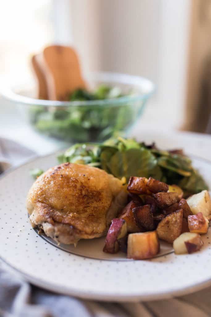 lemon rosemary chicken with crispy potatoes and salad on a white and gray dotted plate. A glass bowl of salad in the background
