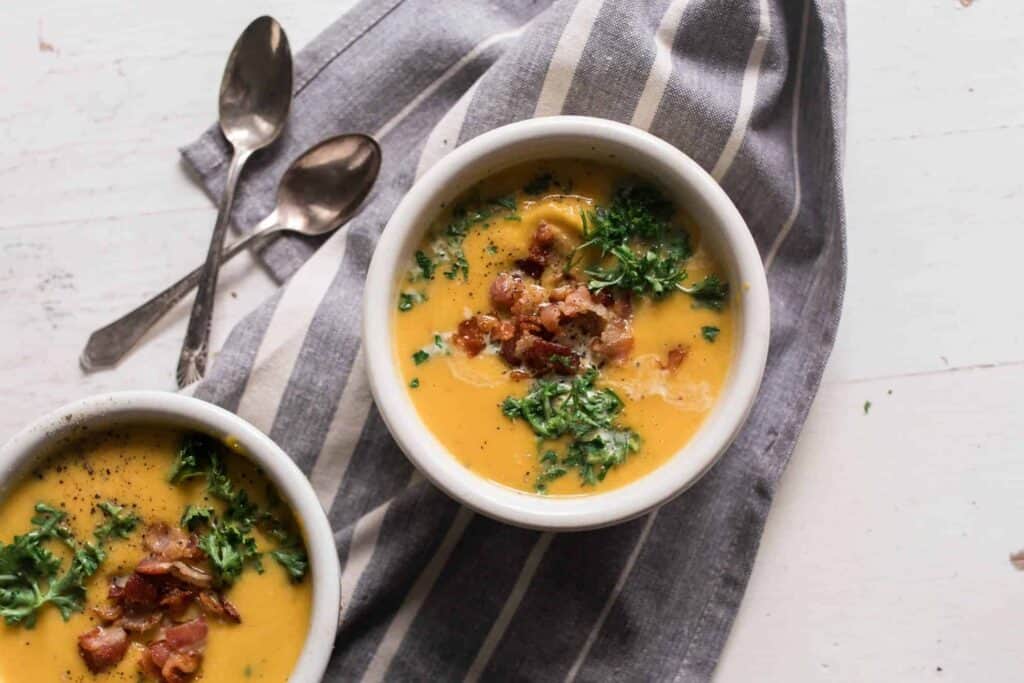 Roasted pumpkin soup with acorn squash in two bowls, topped with fresh herbs and bacon. Bowls lay on top a stripped towel