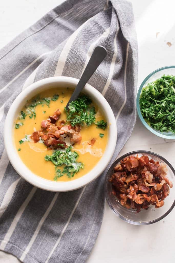 pumpkin soup in a white bowl with parsley and bacon on top a stripped towel with bacon and parsley in jars to the right.