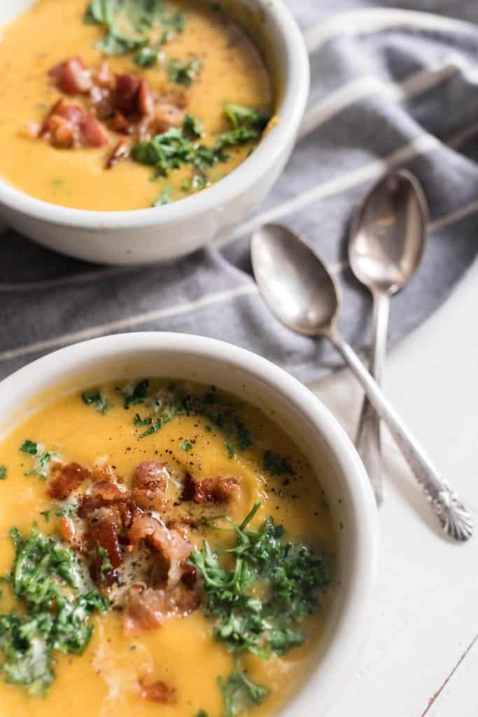 close up photo of a bowl of roasted pumpkin soup topped with bacon and fresh herbs. Antique spoons to the right