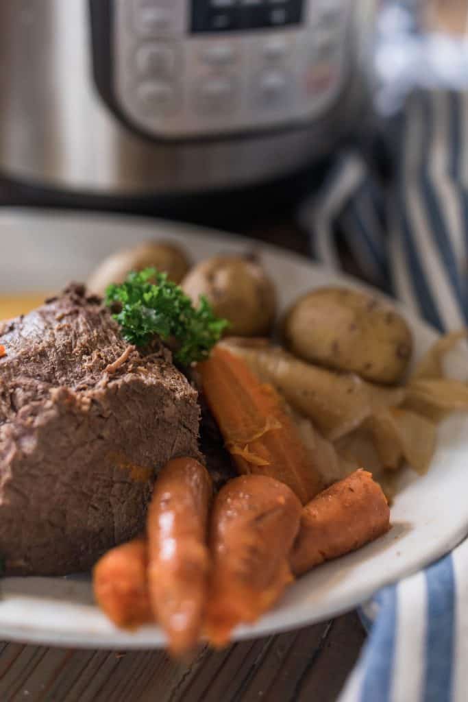 instant pot pot roast with carrots and potatoes on a white plate and garnished with parsley. A blue and white towel to the right of the plate and an Instant Pot behind.