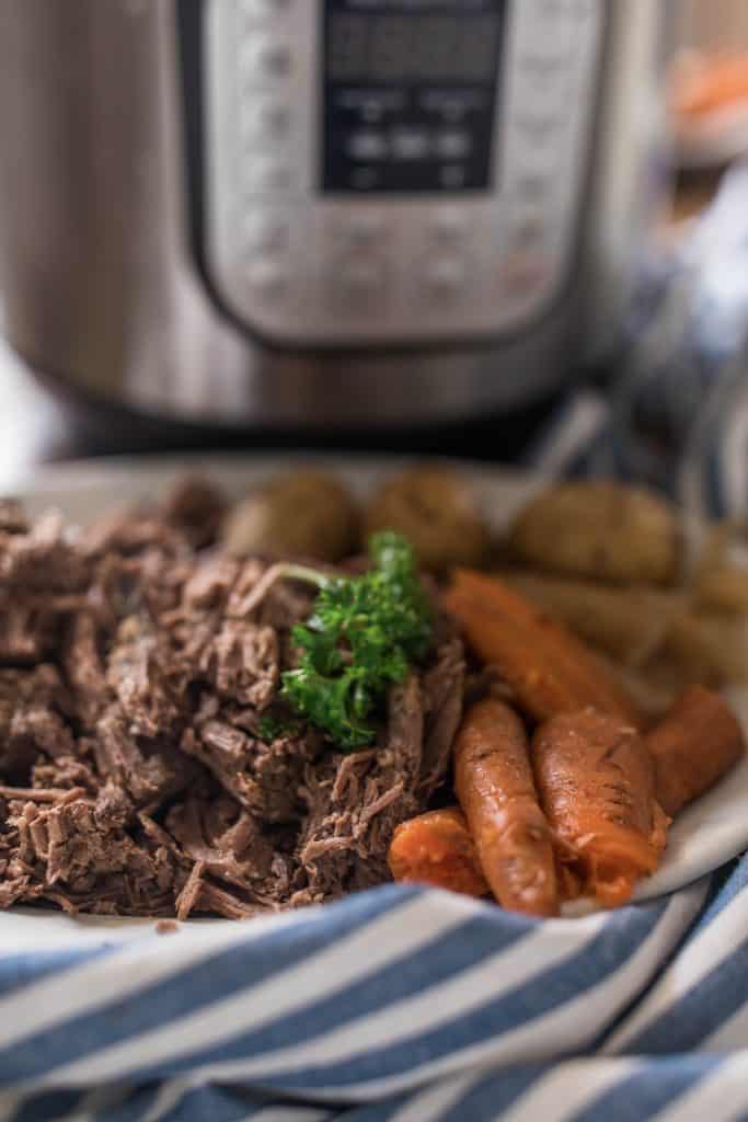 pot roast with carrots and potatoes made on a white plate that is on top a blue and white stripped towel. An instant pot is in the background