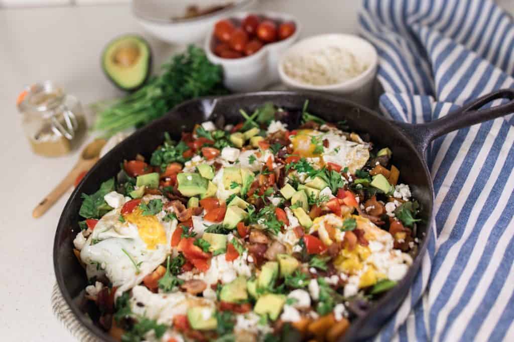 vegetable hash with fresh herbs and eggs in a cast iron skillet. A blue and white stripped towel is to the right