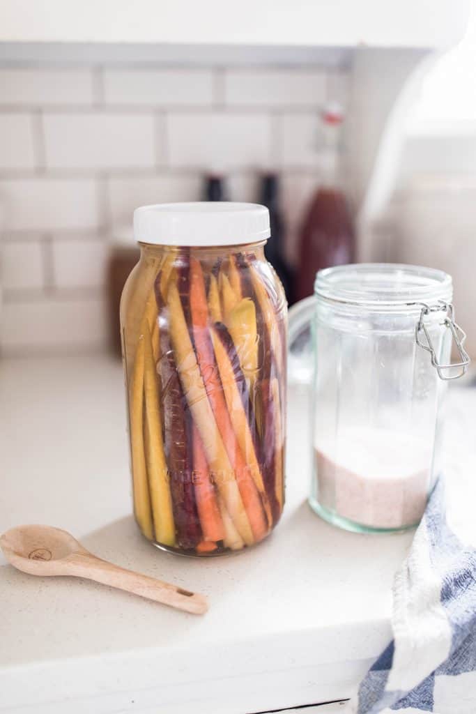 fermented carrots in a half gallon mason jar on a white quarts countertop next to a jar of salt and a wooden spoon