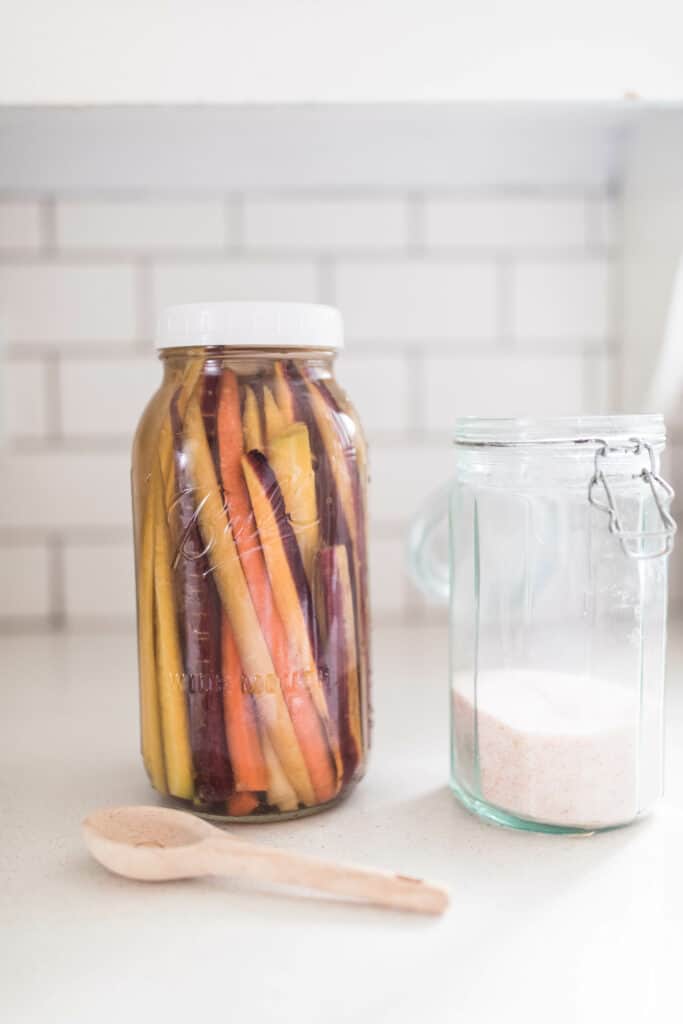 fermented carrots on a white quarts countertop next to a jar or salt and a wooden spoon.