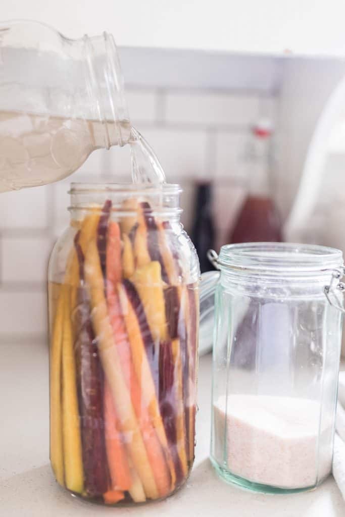 pouring salt water brine over a jar of slice carrots to make fermented carrots