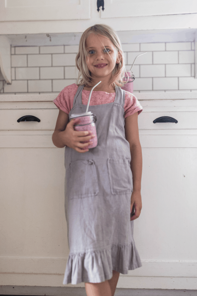 girl with blond hair wearing an apron drinking a berry kefir smoothie in a mason jar