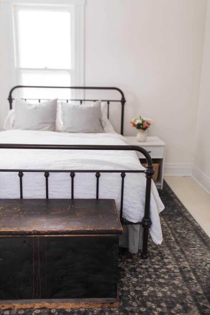 a cozy farmhouse bedroom with a warm rug, large wooden blanket box, iron bed, and farmhouse flowers