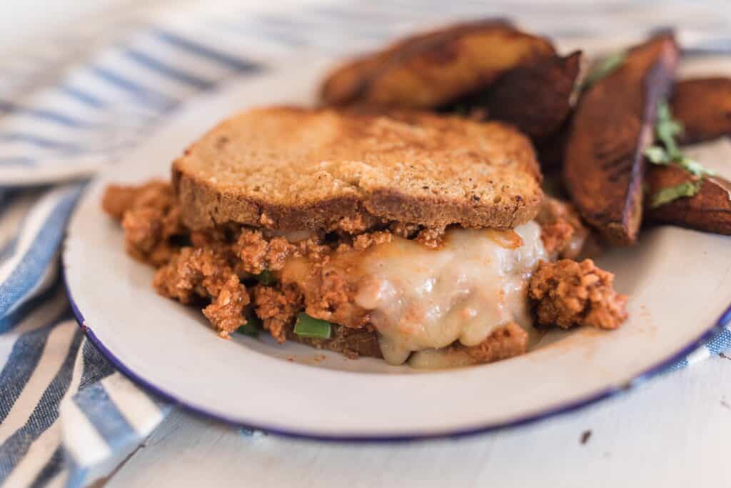 sloppy joes with turkey and cheese on sourdough bread with a side of potato wedges on a white plate on a white wooden table and a blue and white stripped napkin