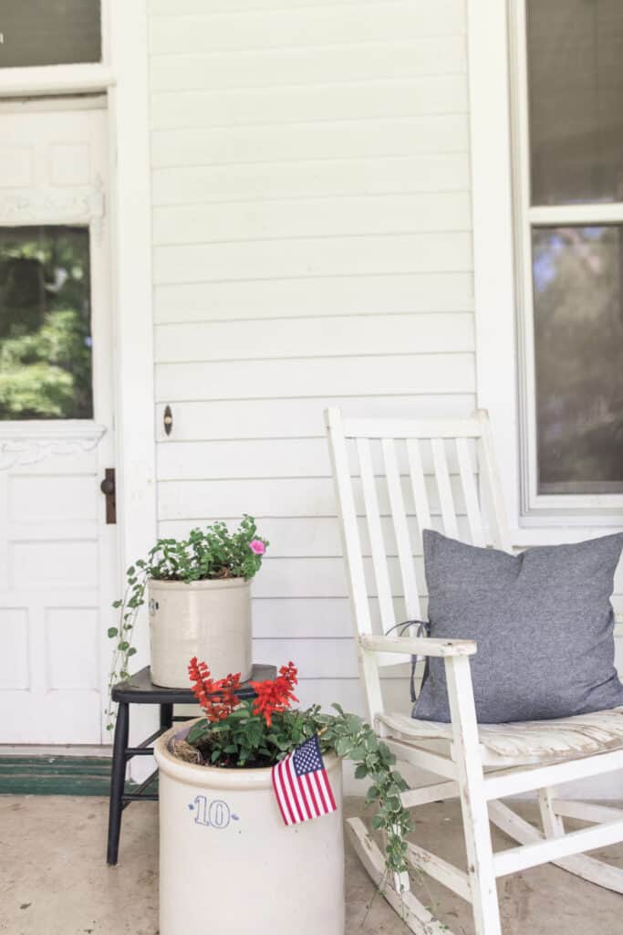 ferns and flowers in crocks and a white chair with blue pillow on a farmhouse front porch