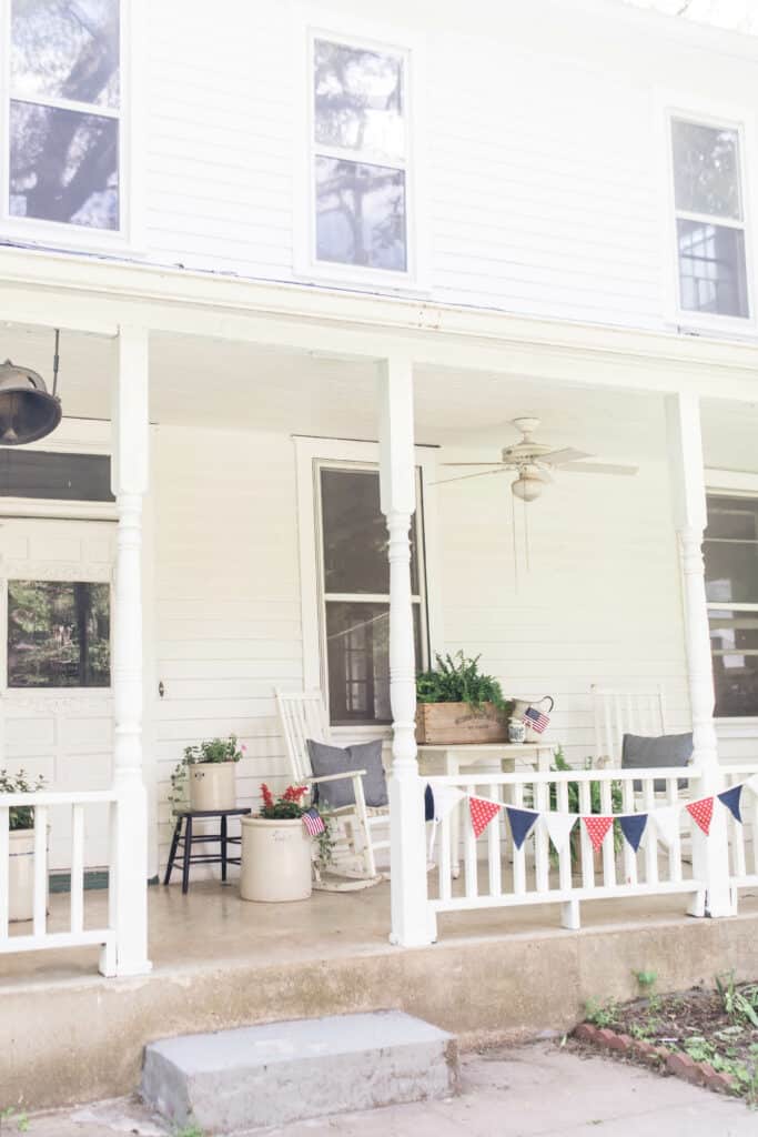 farmhouse summer porch decorated for fourth of july with bunting, crocks full of flowers and ferns and white farmhouse furniture