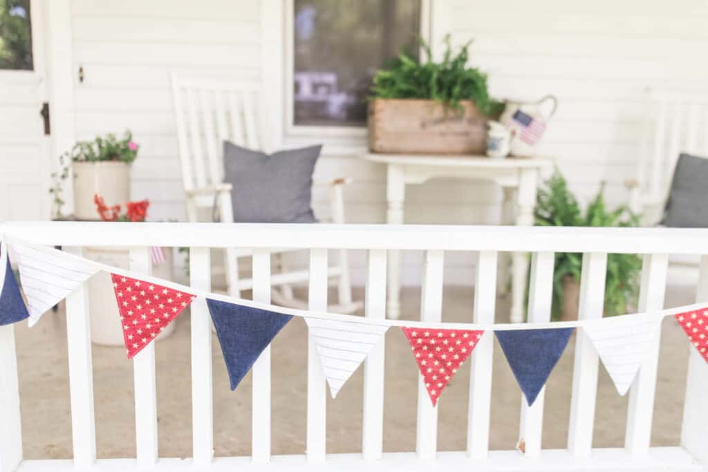 farmhouse front porch with white furniture, blue pillows, forth of July bunting, and plants