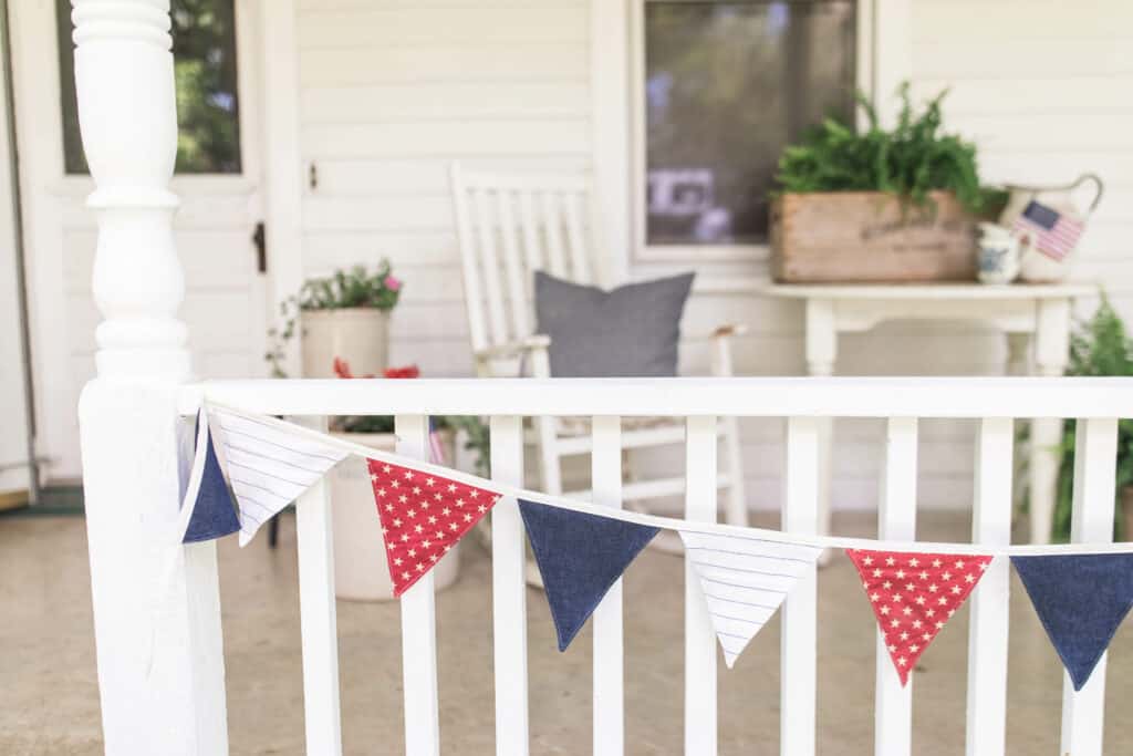 red, white, and blue bunting hanging from a white porch railing with white porch furniture behind it and ferns 