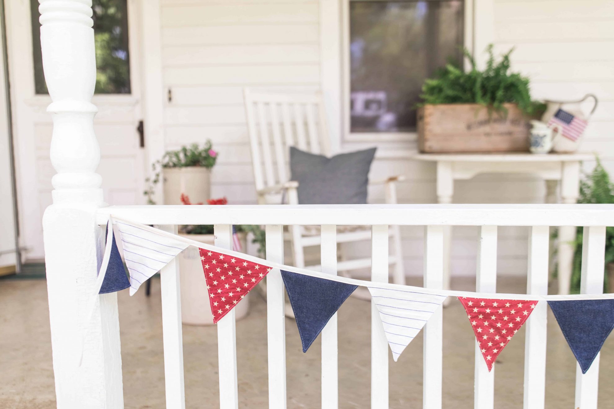 red, white, and blue bunting hanging from a white porch railing with white porch furniture behind it and ferns