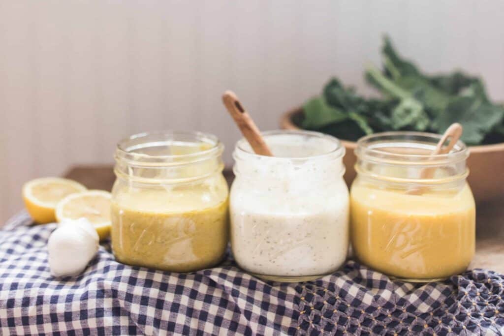 3 homemade dressings: honey mustard, ranch, and lemon vinaigrette dressing in mason jars on white and blue checked towels with a bowl of salad behind them.