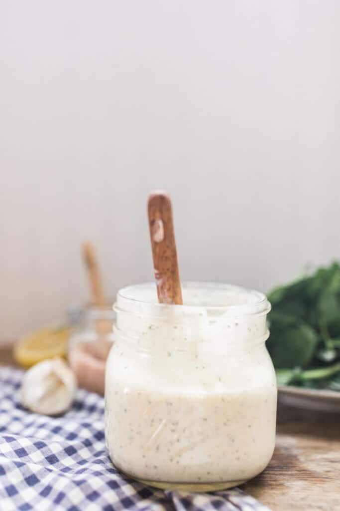 homemade ranch dressing in a mason jar with a wooden spoon in it on top a wood countertop and blue and white checked napkin with greens behind it