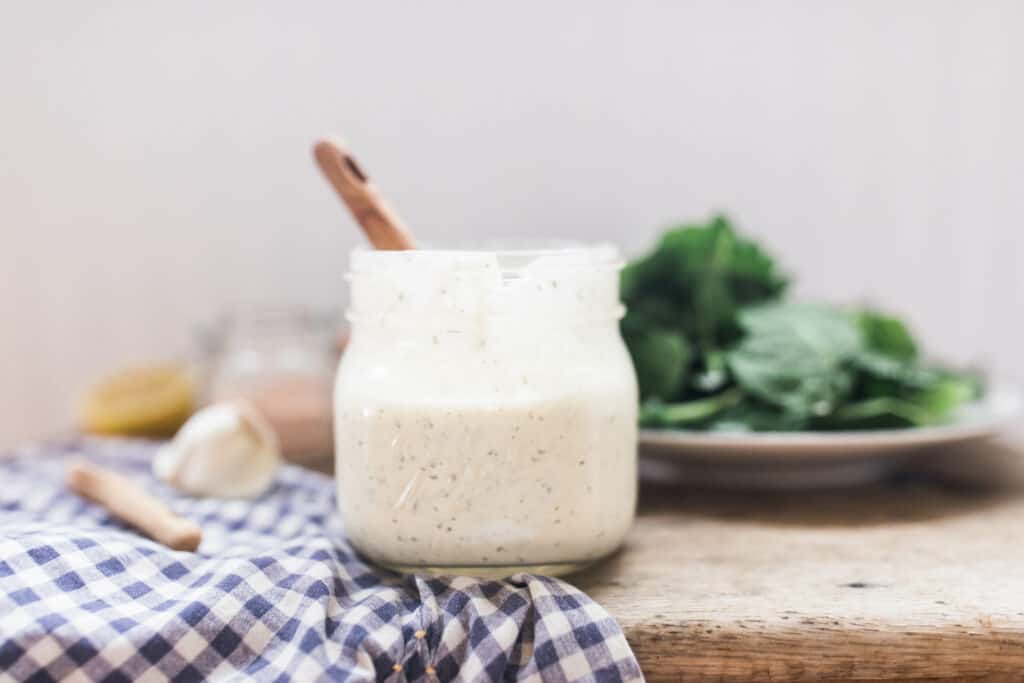 homemade ranch dressing in a mason jar wit wooden spoon on top a blue and white checked towel and a plate of greens behind it