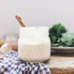 homemade ranch dressing in a mason jar wit wooden spoon on top a blue and white checked towel and a plate of greens behind it