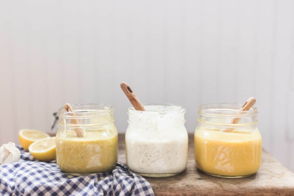 3 homemade salad dressings in glass jars with wooden spoons. On top a white and blue checked towel