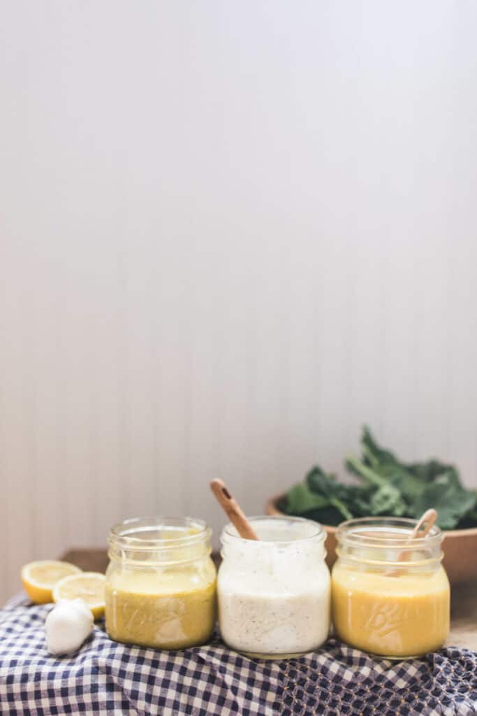 3 homemade salad dressings in glass jars on a wood countertop with a blue and white checked towel and a bowl of salad behind them