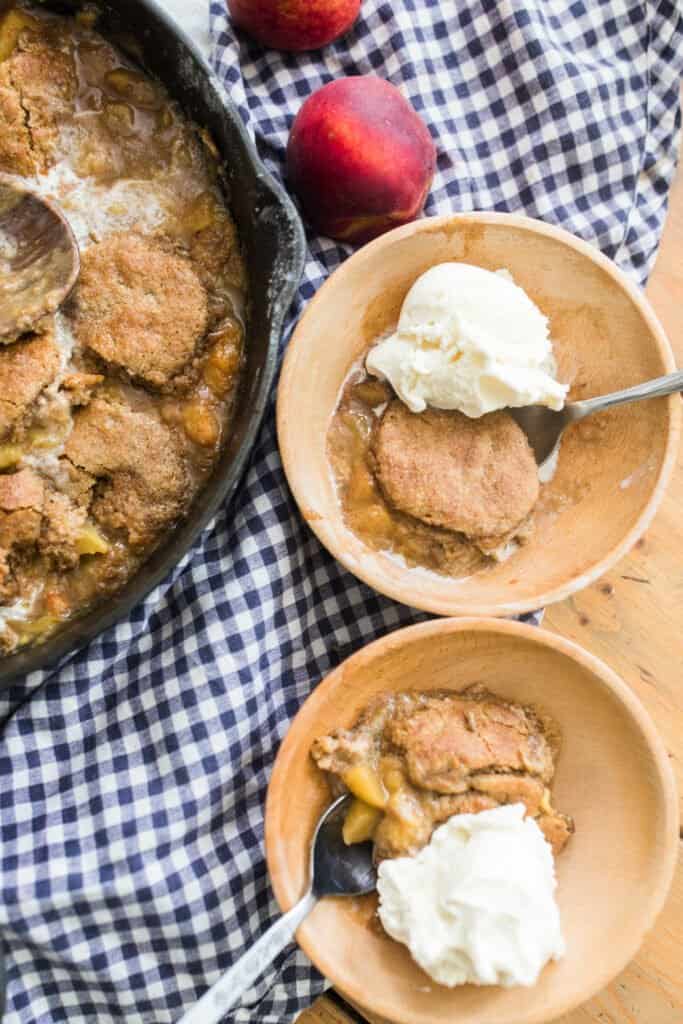 two bowls of peach cobbler in wooden bowls topped with ice cream on a blue and white checked towel and a cast iron skillet with remaining cobbler