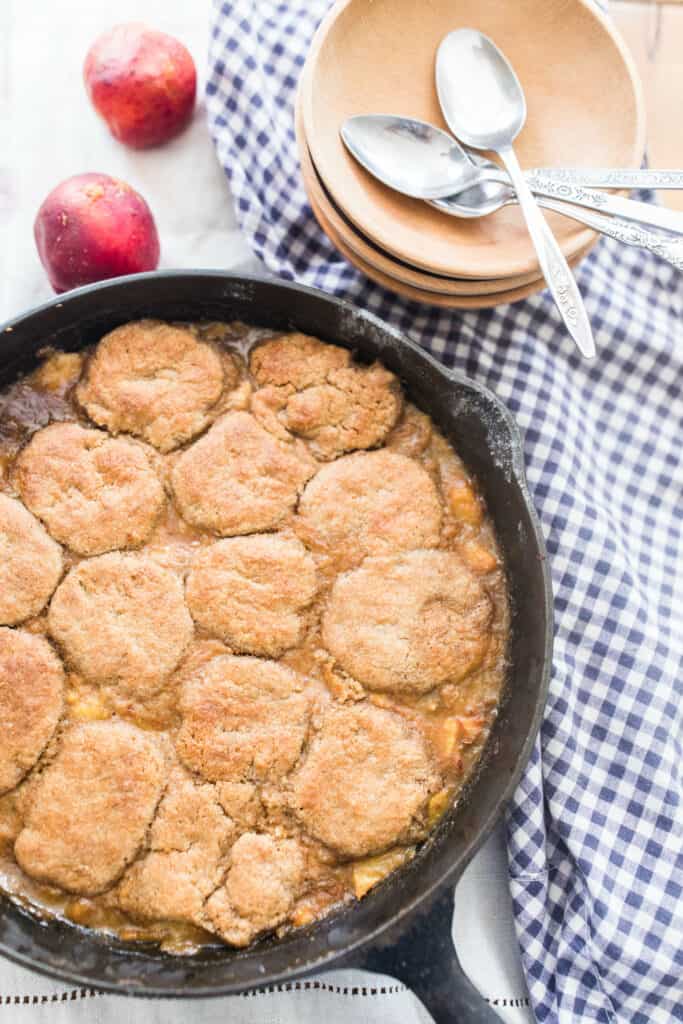 sourdough peach cobbler in a cast iron skillet on a blue and white towel with wooden bowls behind it